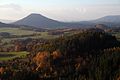 Farbfotografie in der Vogelperspektive von einem Tal mit mehreren Hügeln. Die Hügel sind mit herbstlichen Bäumen bestückt und dazwischen befinden sich Wiesen. Im Hintergrund sind größere Berge zu erkennen.