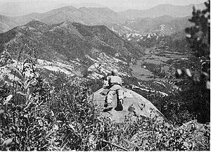 A man lies on a cliff overlooking a large hill complex