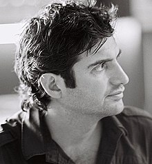 Man with open collared shirt and tousled hair looks upwards