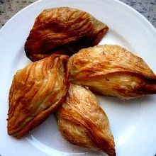 La photographie couleur présente une assiette blanche garnie de quatre pastizzi rôtis. Les stries apparentes montrent bien qu'il s'agit de pâte feuilletée.