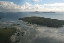 Two dark green areas of land are separated from one another by a shallow and narrow strait. There are small islands in the distance under white clouds.