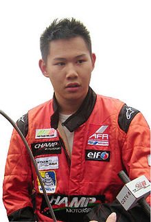 Photographie d'un homme asiatique, vu de face, portant une combinaison orange, devant un micro.