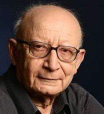 Colour publicity shot of an elderly, bald man with spectacles and a black shirt.