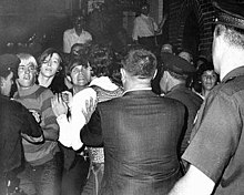 A black and white photograph showing the backs of three uniformed police officers and a man with short-cropped hair in a suit pushing back a crowd of young men with longer hair dressed in jeans and contemporary clothing for the late 1960s, arguing and defying the police; other people in the background on a stoop are watching