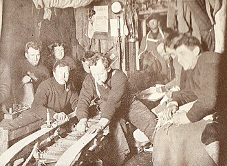 A group of men in woollen jerseys, several smoking pipes, are watching repair work on a sledge. They are in a confined area, with equipment and spare clothing adorning the walls