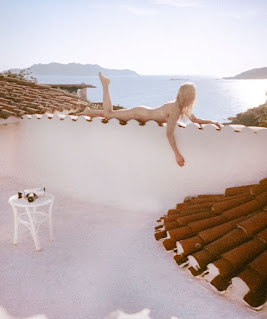 Fotografía. Azotea de una casa de estilo mediterráneo, con suelo blanco y muro blanco. Tejado de tejas marrones. Sobre este, una chica joven tumbada bocabajo, desnuda, de piel clara y pelo rubio. Mirando hacia el mar.