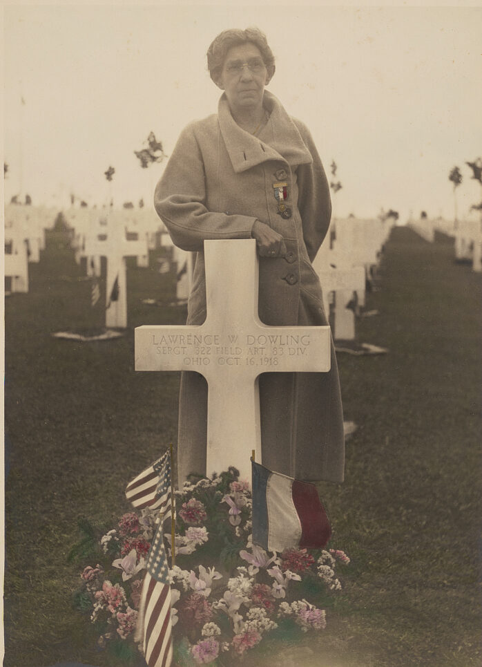 Woman in hat and coat standing infront of gravestone shaped like a cross