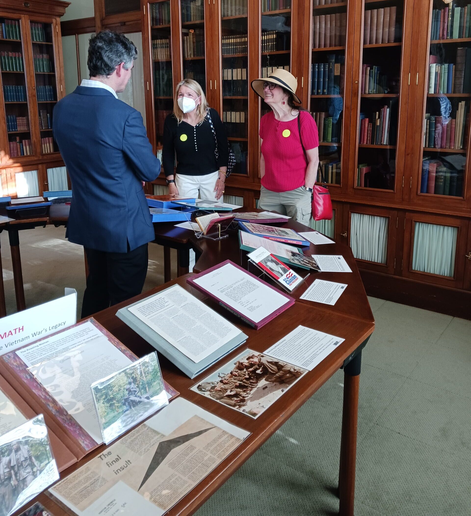 Collection items in foreground, historian speaking with two visitors in background, with wooden bookshelves against wall