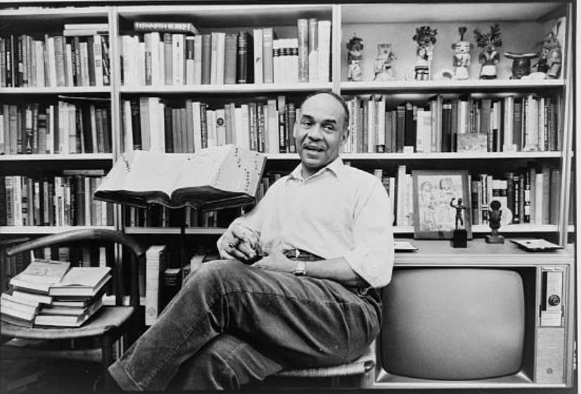 Ralph Ellison seated cross-legged in front of a wall of books, television set at his left.