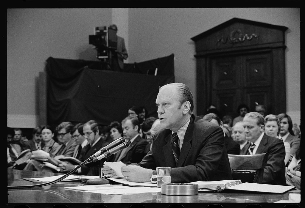 Monochrome image of Ford at table in committee hearing room, speaking into microphone