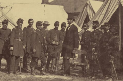 Monochrome photograph of Lincoln standing with hand on chair, uniformed Union soldiers standing at left and right, with military tents in background