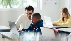 Male teacher helps a young boy with computer-based learning in a classroom setting. Child tutor providing a lesson in an elementary school, with a focus on coding and basic digital literacy.