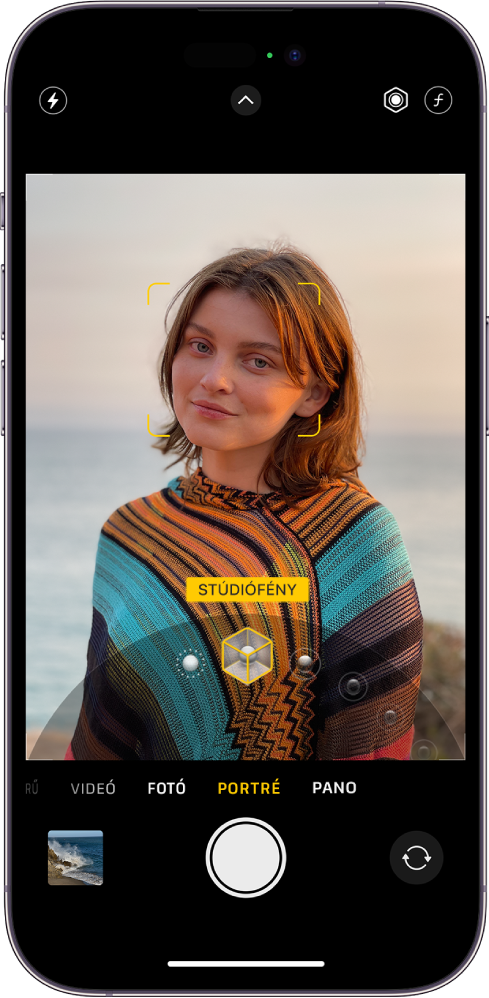A kamera képernyője Portré módban; a téma élesen látható a keresőben, miközben a háttér elmosódott. A keret alsó részén meg van nyitva a portrémegvilágítás kiválasztásához használható tárcsa, amelyen a Stúdiófény van kiválasztva. A képernyő bal felső részén a Vaku gomb látható, középen felül a Kameravezérlők gomb, a jobb felső részen pedig a portrémegvilágítás intenzitásának és a mélység vezérlésének beállítására szolgáló gombok jelennek meg. A képernyő alján balról jobbra haladva a Fotó- és videonéző gomb, a Kép készítése gomb, illetve a Kameraválasztó hátsó gomb található.
