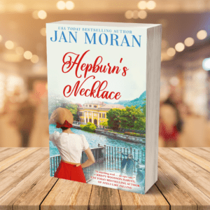 Hepburn's Necklace by Jan Moran Book Cover, a scene overlooking Lake Como, Italy, with lake, villa, Vespa, and Audrey Hepburn-esque woman in the foreground. Includes a quote from author Kristy Woodson Harvey.