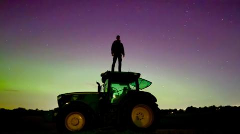 A man stands on top of a tractor with the Northern Lights in the background in Frieth, Buckinghamshire