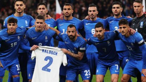 Greece players line up behind a shirt bearing George Baldock's name before the match against England