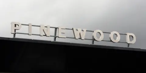 The front gate of Pinewood studios in Iver Heath. The word Pinewood has been fastened to a metal frame. They stand out against a grey sky. 