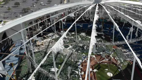 The roof of a stadium from above after being ripped off with the pitch visible below