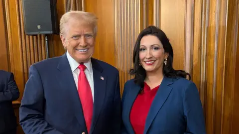 Donald Trump gives a thumbs up as he meets Deputy First Minister Emma Little-Pengelly. She is wearing a red dress with a blue blazer. The pair are standing in a wooden panelled room.