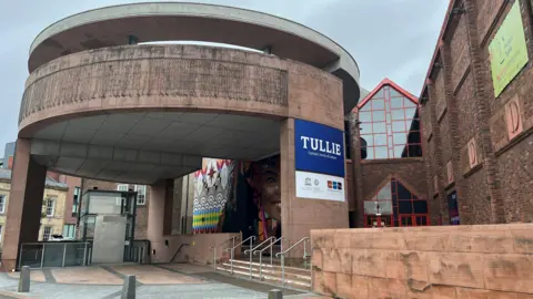 A general view of the entrance to Tullie museum in Carlisle. The entrance is formed of a circular building with a colourful mural in the background.