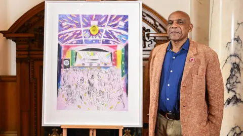 British artist Denzil Forrester - a man smiling, wearing a navy buttoned shirt, a brown tweed suit jacket and light brown trousers, standing to the right of his artwork depicting a reggae festival in Cornwall.