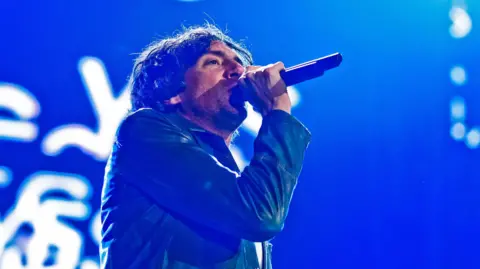 Gary Lightbody sings into a microphone on stage wearing a blue jacket. Behind him is a blue screen with white letters.