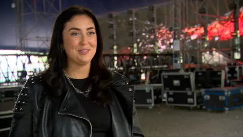 DJ K.A.D in a black leather jacket. She has long black hair and is smiling at the camera in front of a stage. 