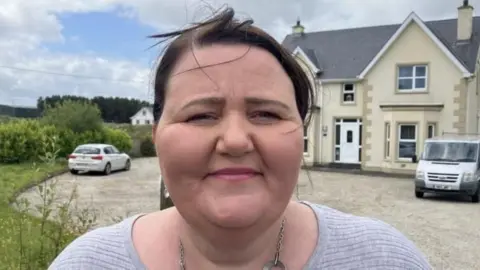 A woman stands outside her home. two vehicles are parked behind her. She is wearing a silver necklace, has brown hair pulled back and is wearing a grey top