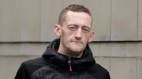 A man with short cropped hair is looking into the camera. He is wearing a black zip up jacket and is standing in front of a stone wall. 