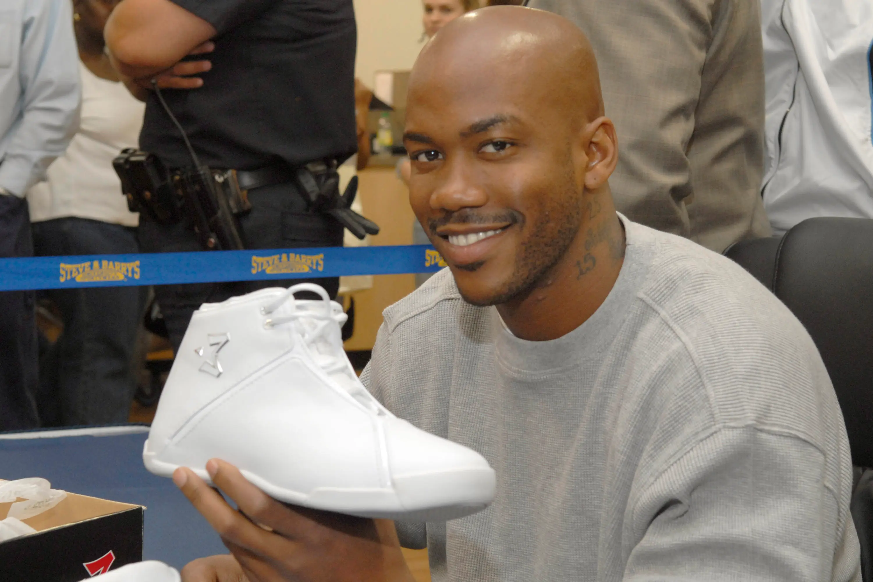 A man holding a white athletic shoe, smiling at an event with people in the background.