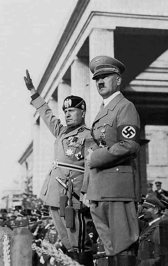 Hitler og Mussolini står oppe på et podium. Mussolini holder den ene armen løftet til en fascistisk hilsen. Folkemengde i bakgrunnen.