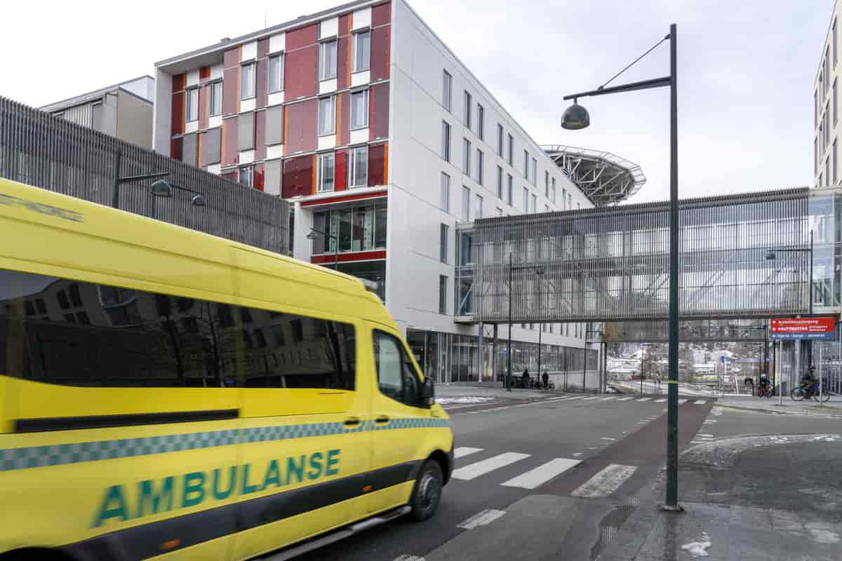 Foto fra sykehusområdet i Trondheim med en gul ambulanse i fart. På siden av bilen står det "AMBULANSE", og farten gjør at den er litt uskarp. I bakgrunnen er det et stort, hvitt sykehusbygg med vinduer og fargerike paneler i rødt og oransje på fasaden. På taket synes litt av en landingsplattform for helikopter. Mellom bygningene går en gangbro av metall og glass, som forbinder ulike deler av sykehuset. 