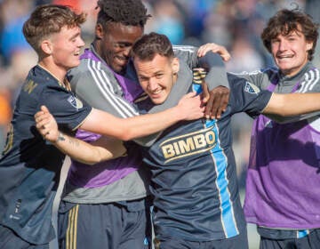 Soccer players surround and hug one soccer player who is smiling.