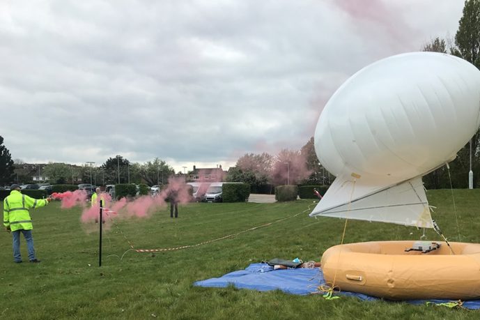 A white balloon and a white kite float above a yellow, ring-shaped inflatable, which is sitting on the ground on a blue tarpaulin. The kite and balloon are tethered to a pole. A person in a hi-vis vest stands to theleft holding a flare that is emitting pink smoke.