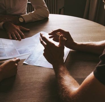 Hendene til tre personer som sitter rundt et bord. Hendene gestikulerer over papirer.