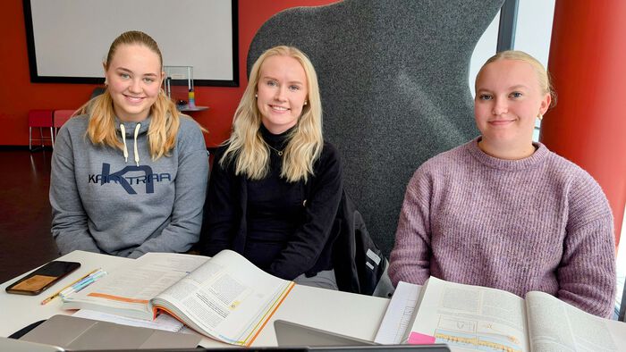 Tre unge damer sitter i biblioteket med bøker oppslått på et bord foran seg.