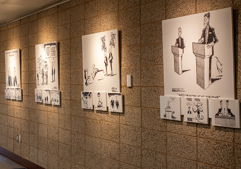 Wall-mounted display of black and white political cartoons in an indoor gallery.