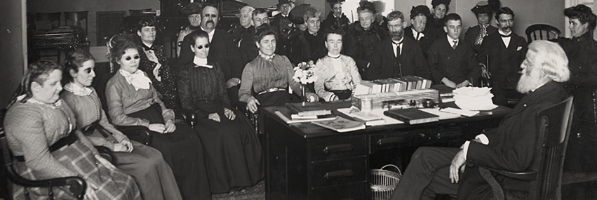 25-30 men and women sit around a desk with books on it. Some wear black glasses and others’ eyes are shut. An older man leans back in his chair behind the desk facing away from the camera.