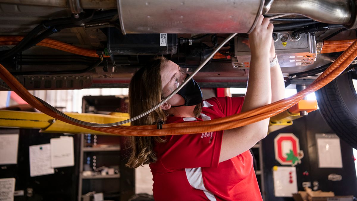 A researcher wearing a mask works on the undercarriage of an automobile.