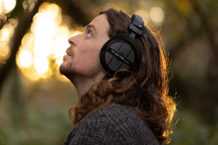 Man with long hair listening to headphones outdoors at sunset.