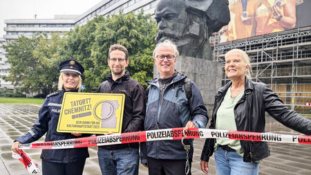 Zwei Männer und zwei Frauen stehen vor dem Chemnitzer Karl-Marx-Monument und halten ein Polizeiabsperrband in den Händen. 