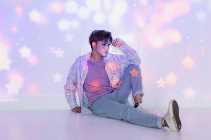 A K-pop star in a purple pastel-themed outfit sits in front of a starry backdrop on a wall.